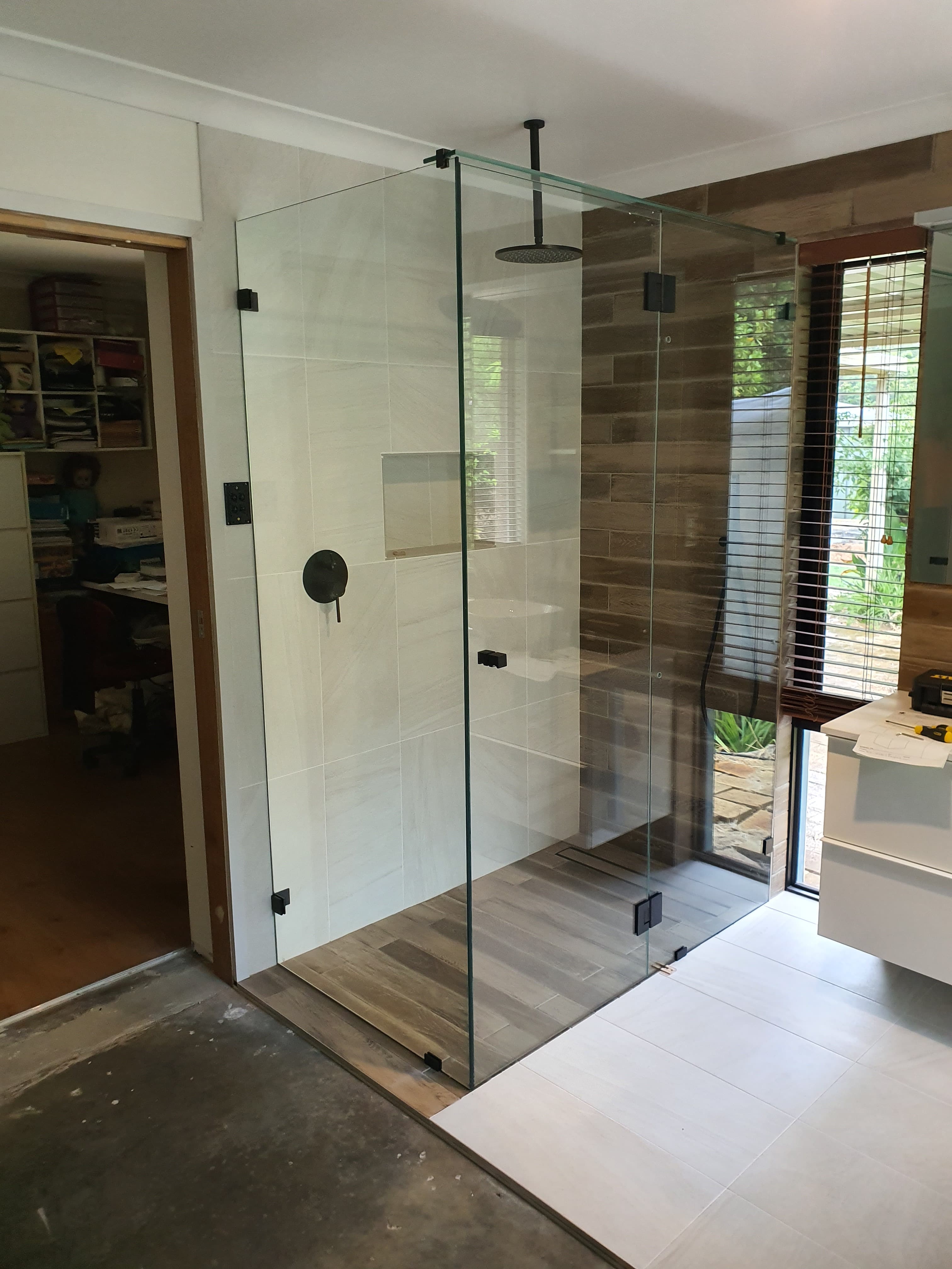 Modern frameless glass shower featuring matte black hardware, white wall tiles, and wood-look floor tiling.