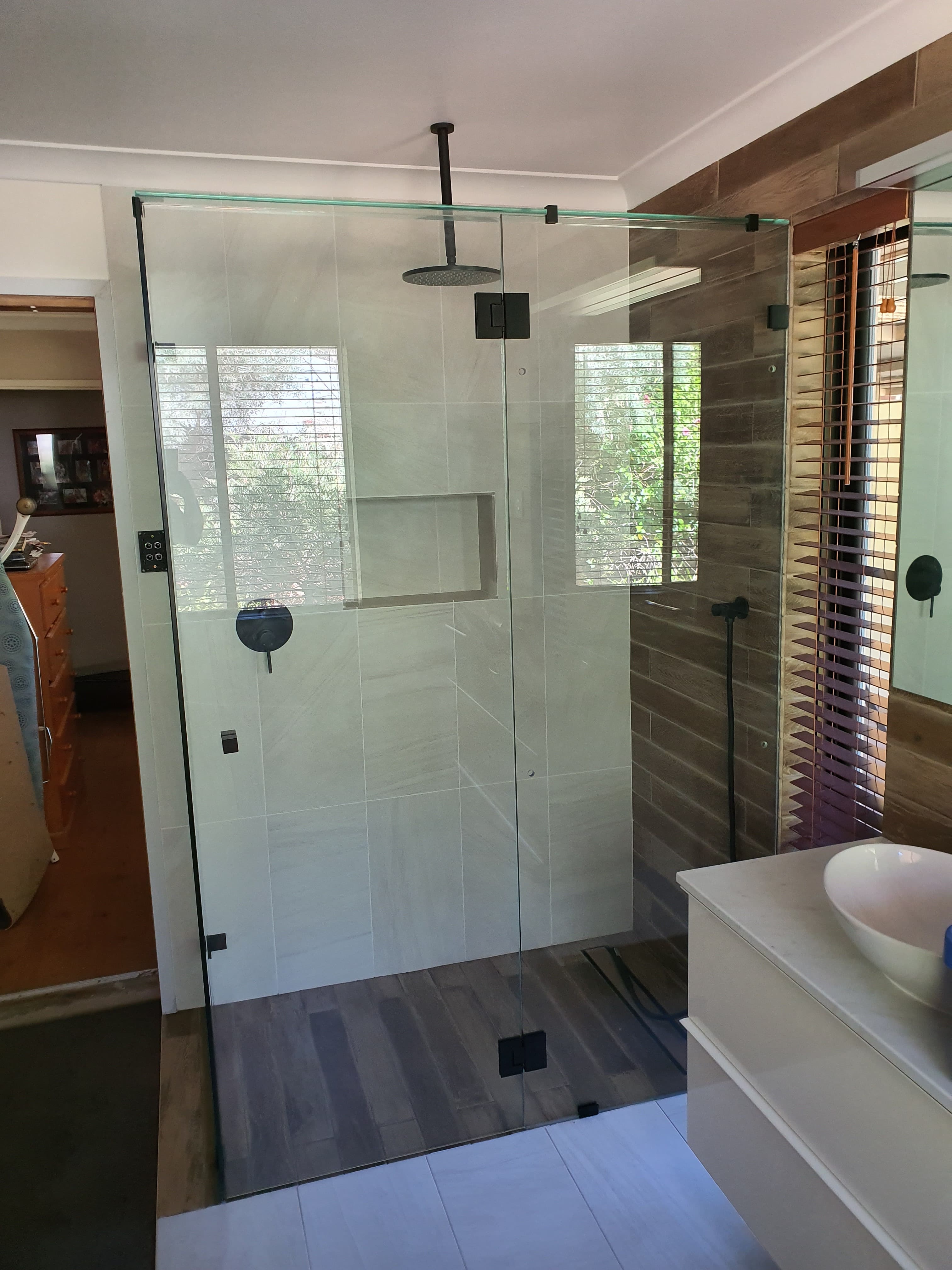 Modern walk-in shower with frameless glass, black fixtures, light gray tiles, and wood-look flooring.