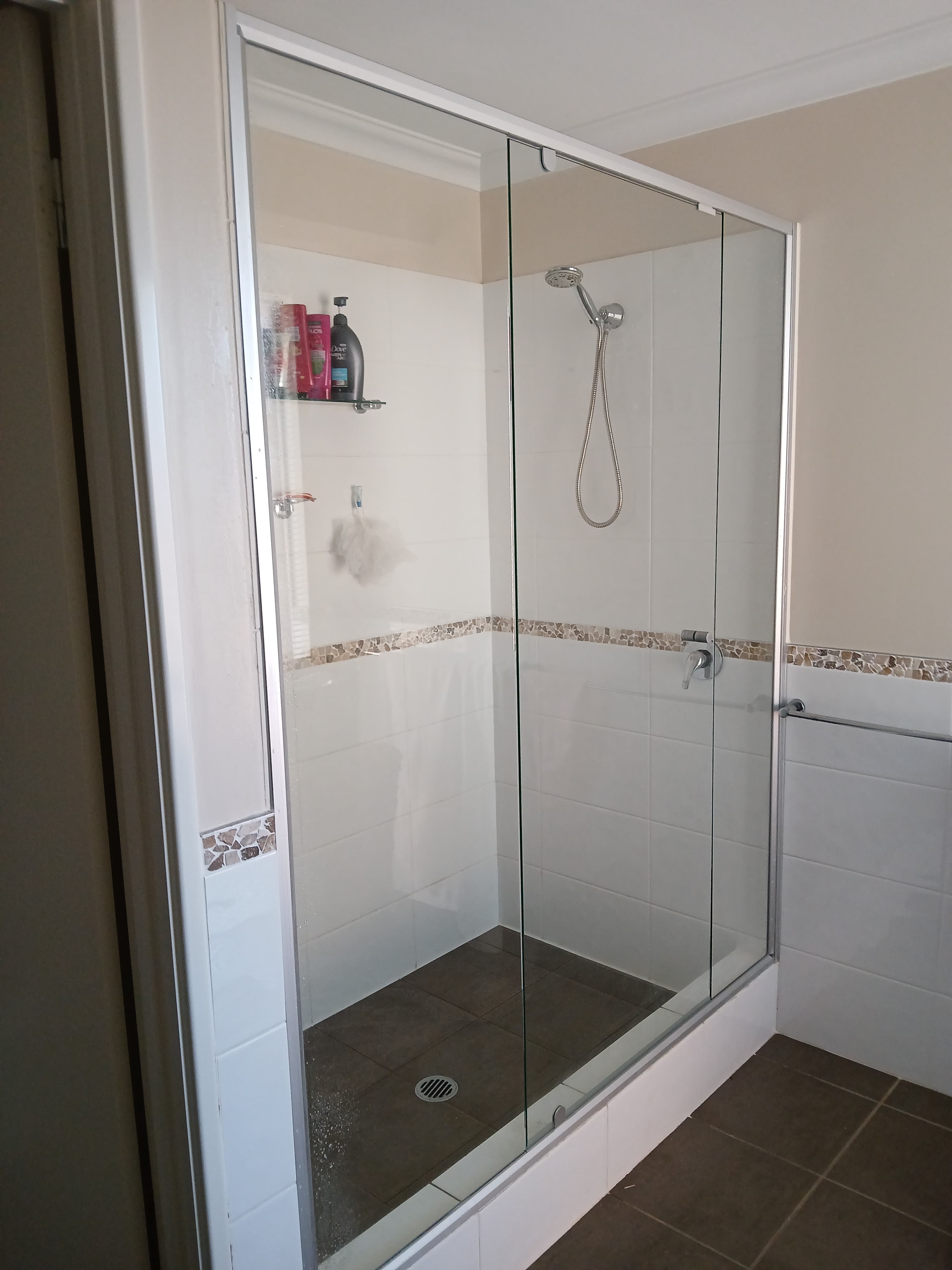 Glass shower enclosure with white tiled walls, mosaic border, chrome fixtures, and dark floor.