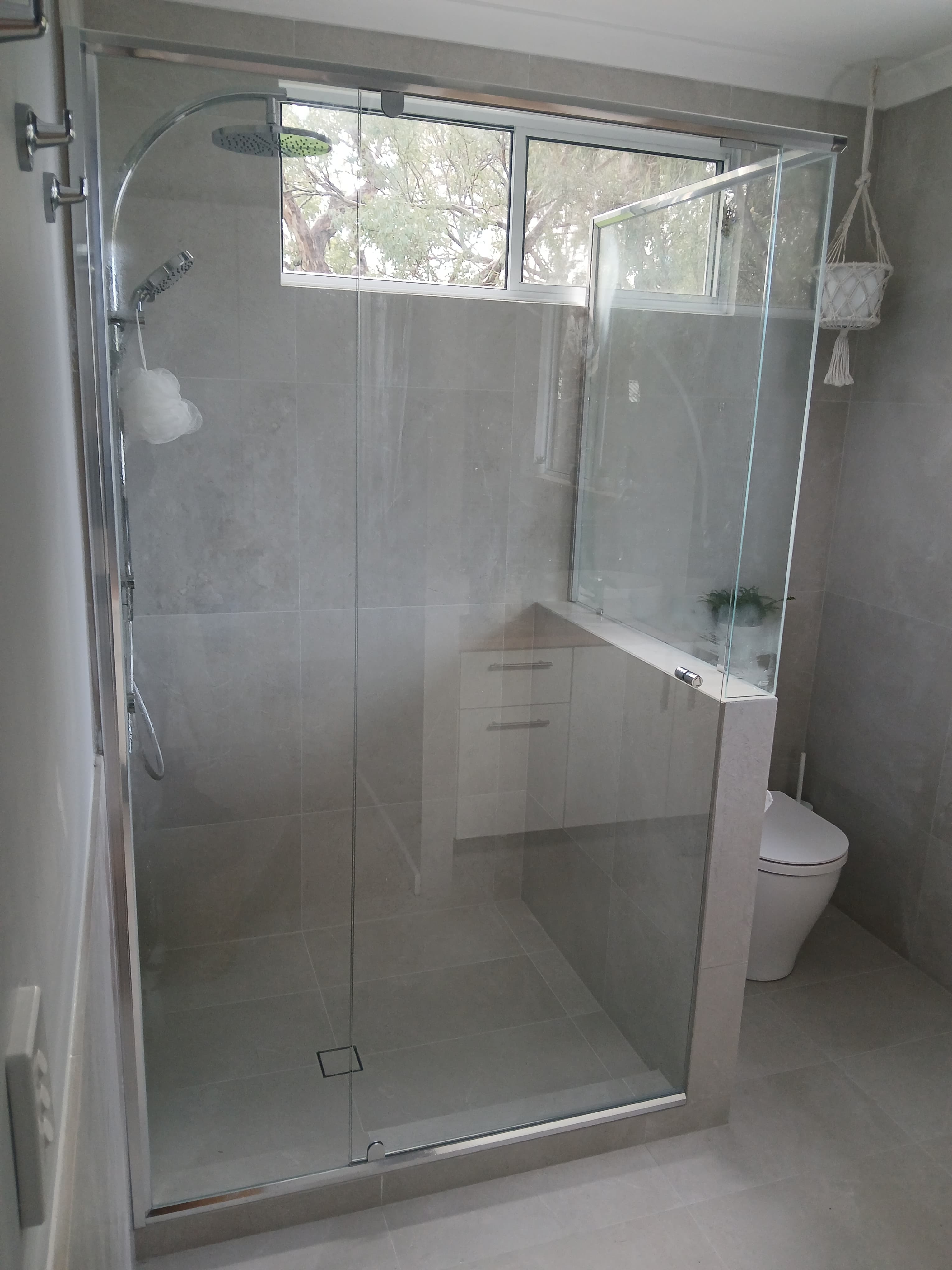 Walk-in glass shower with chrome fixtures, grey tiled walls, and a window overlooking green trees.