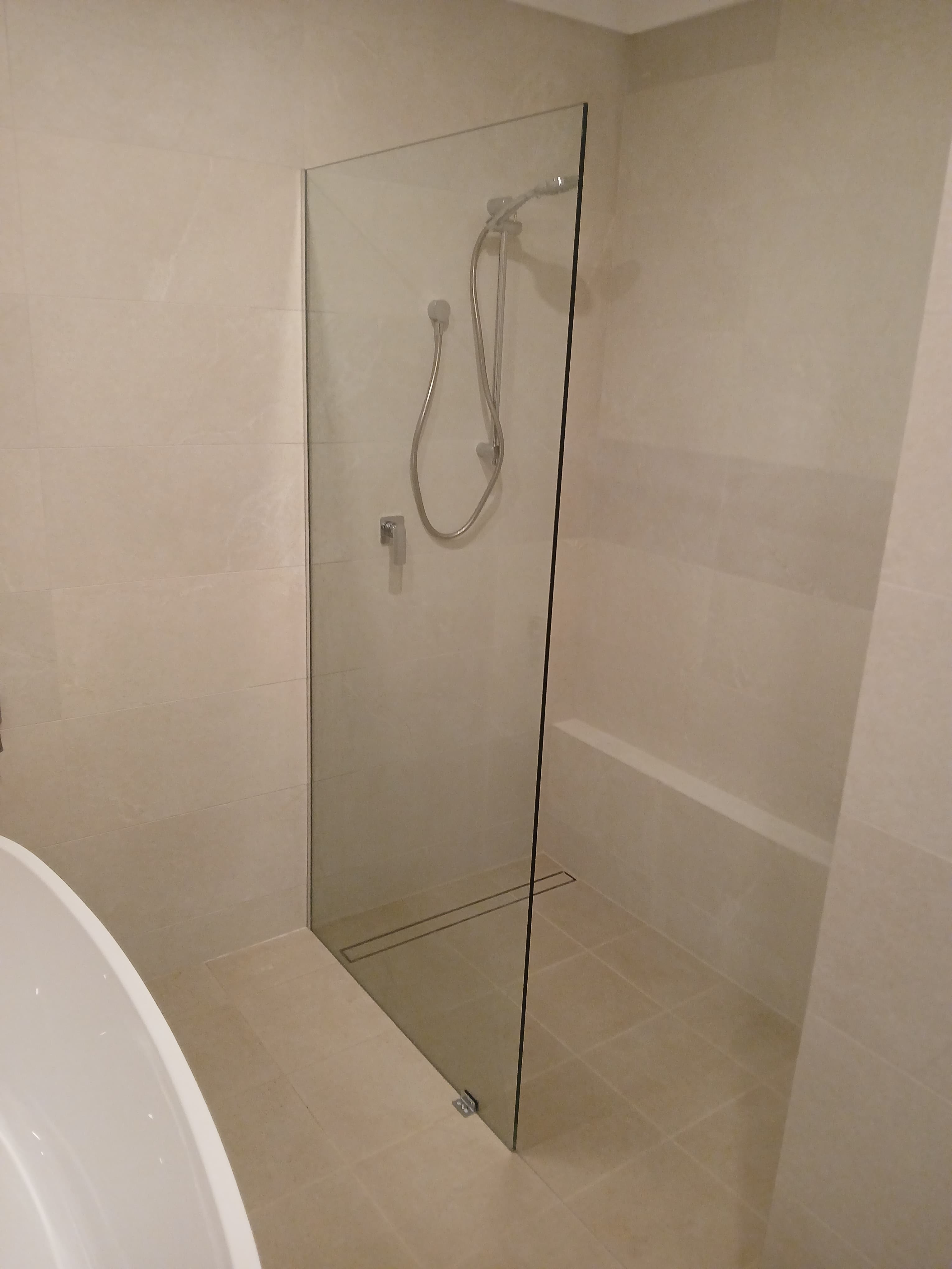 Modern walk-in shower with frameless glass panel, handheld shower head, and beige tiled walls.