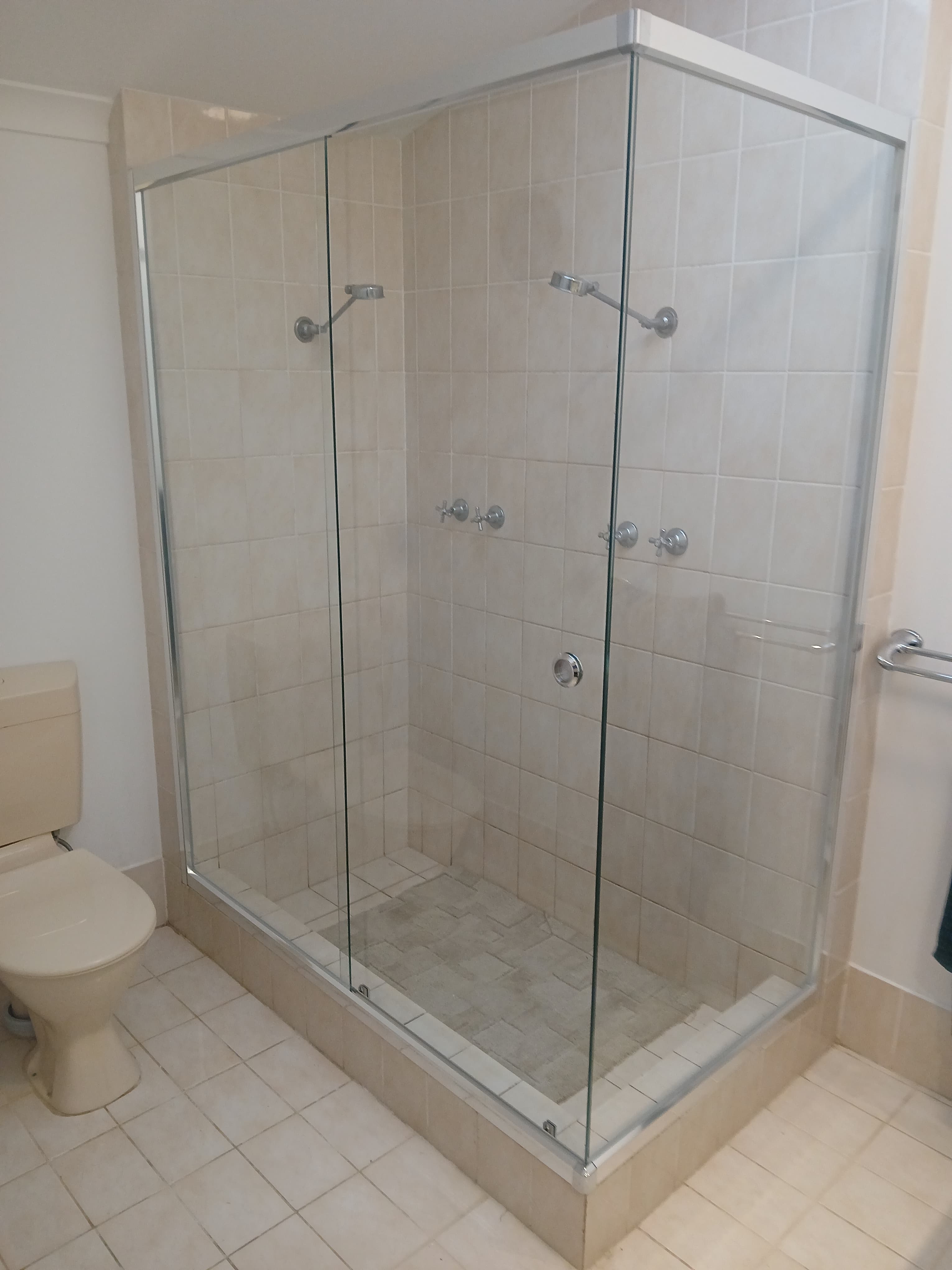 Large glass-enclosed walk-in shower with two shower heads and beige tiles in a neutral bathroom.