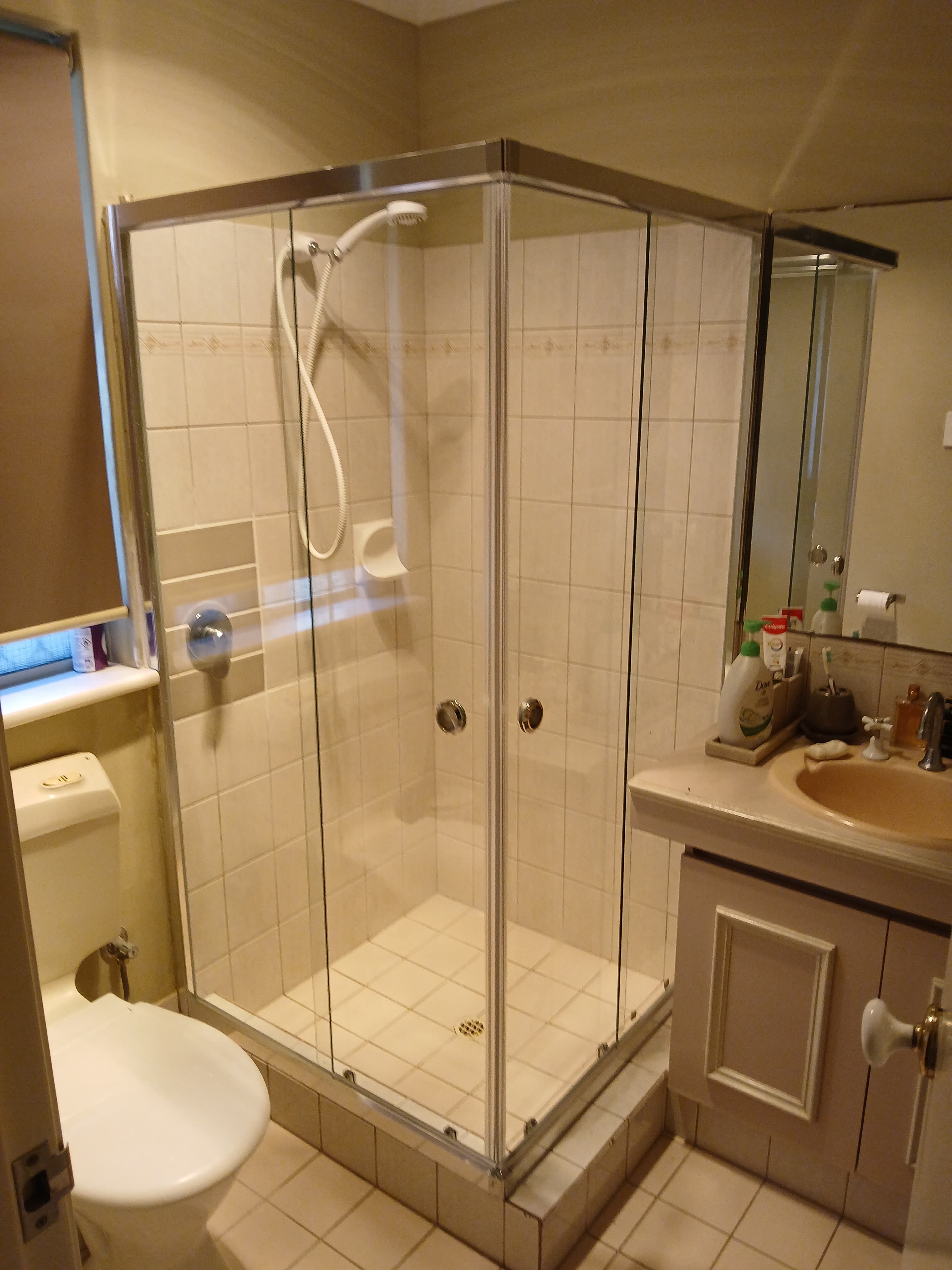Glass-enclosed corner shower, white toilet, and beige vanity in a neutral-toned tiled bathroom.