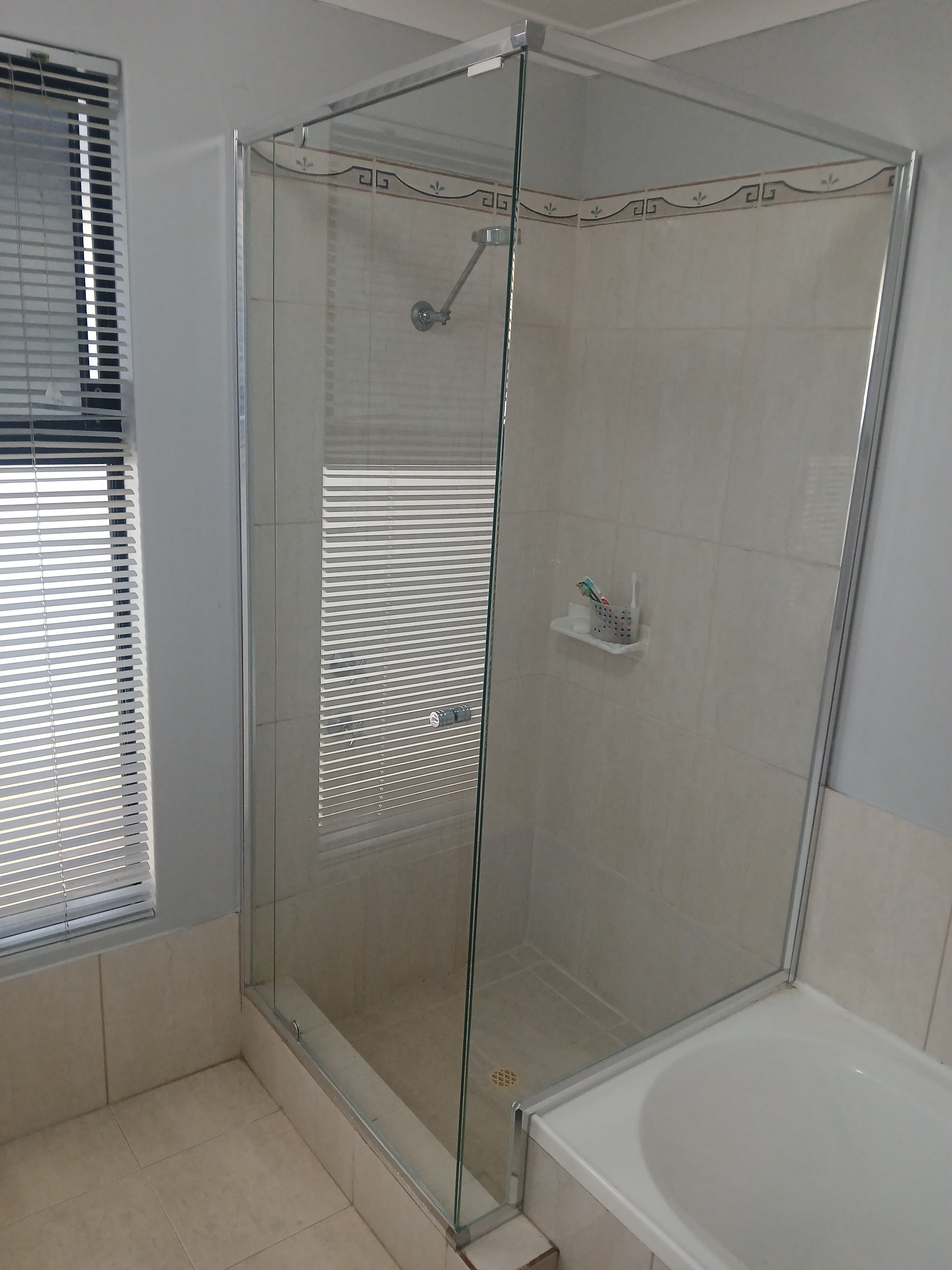 Glass shower enclosure with beige tiles, silver fixtures, and a window with white blinds.