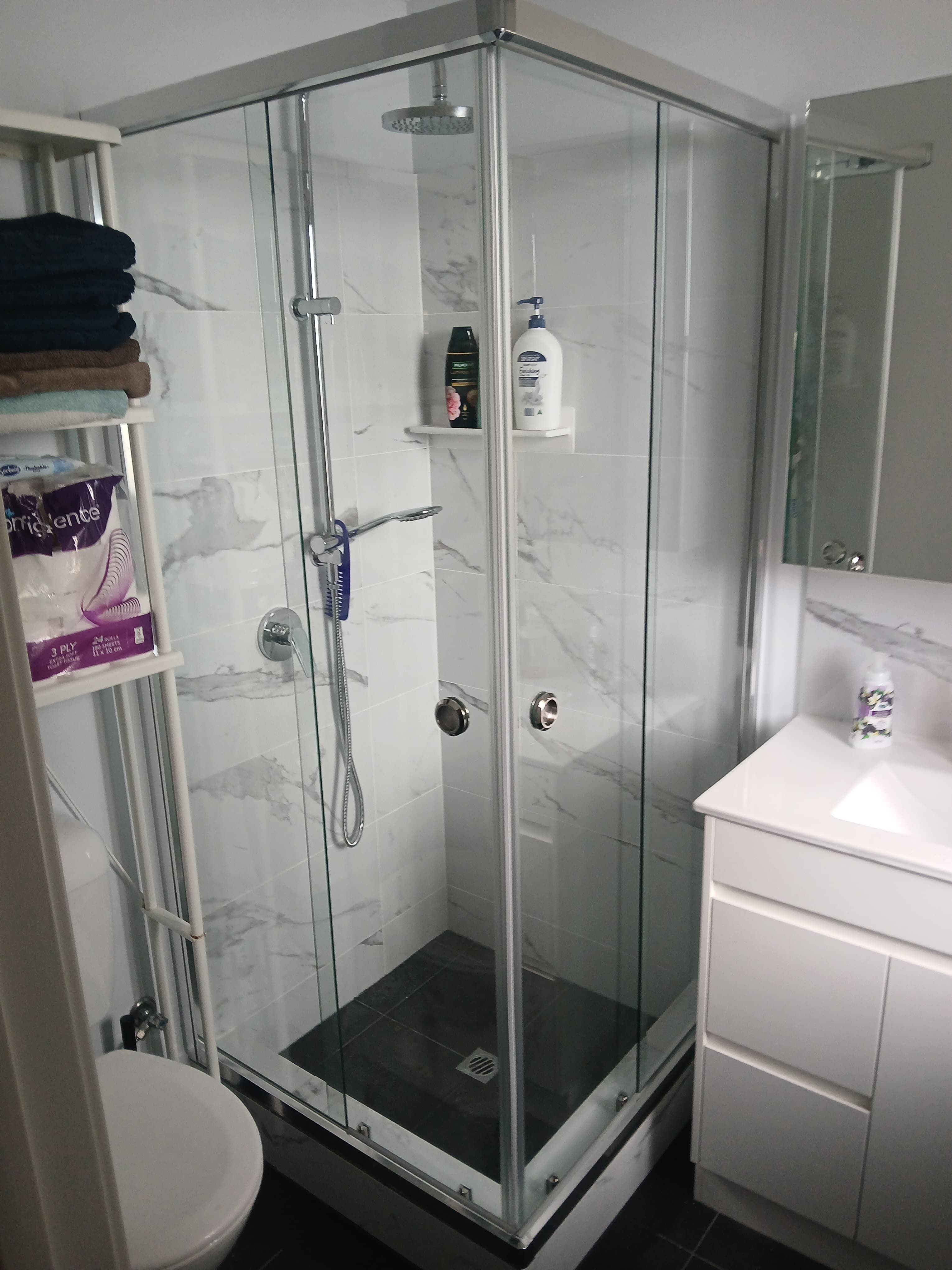 Modern bathroom featuring a glass shower with marble tiles, white vanity, and storage shelving.