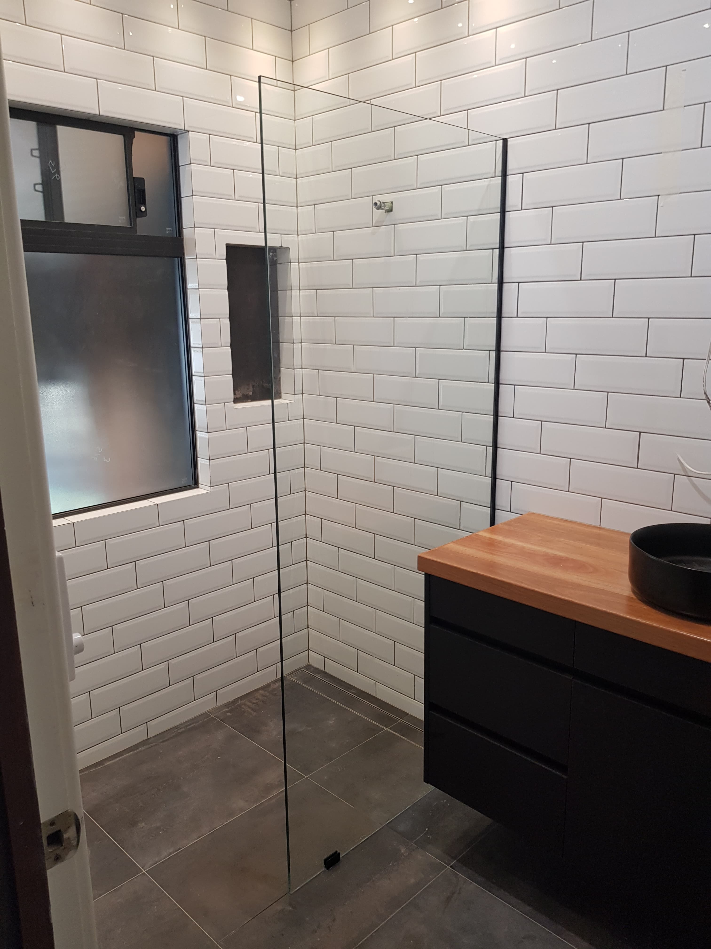 Modern bathroom featuring white subway tiles, glass shower screen, and black vanity with wood countertop.