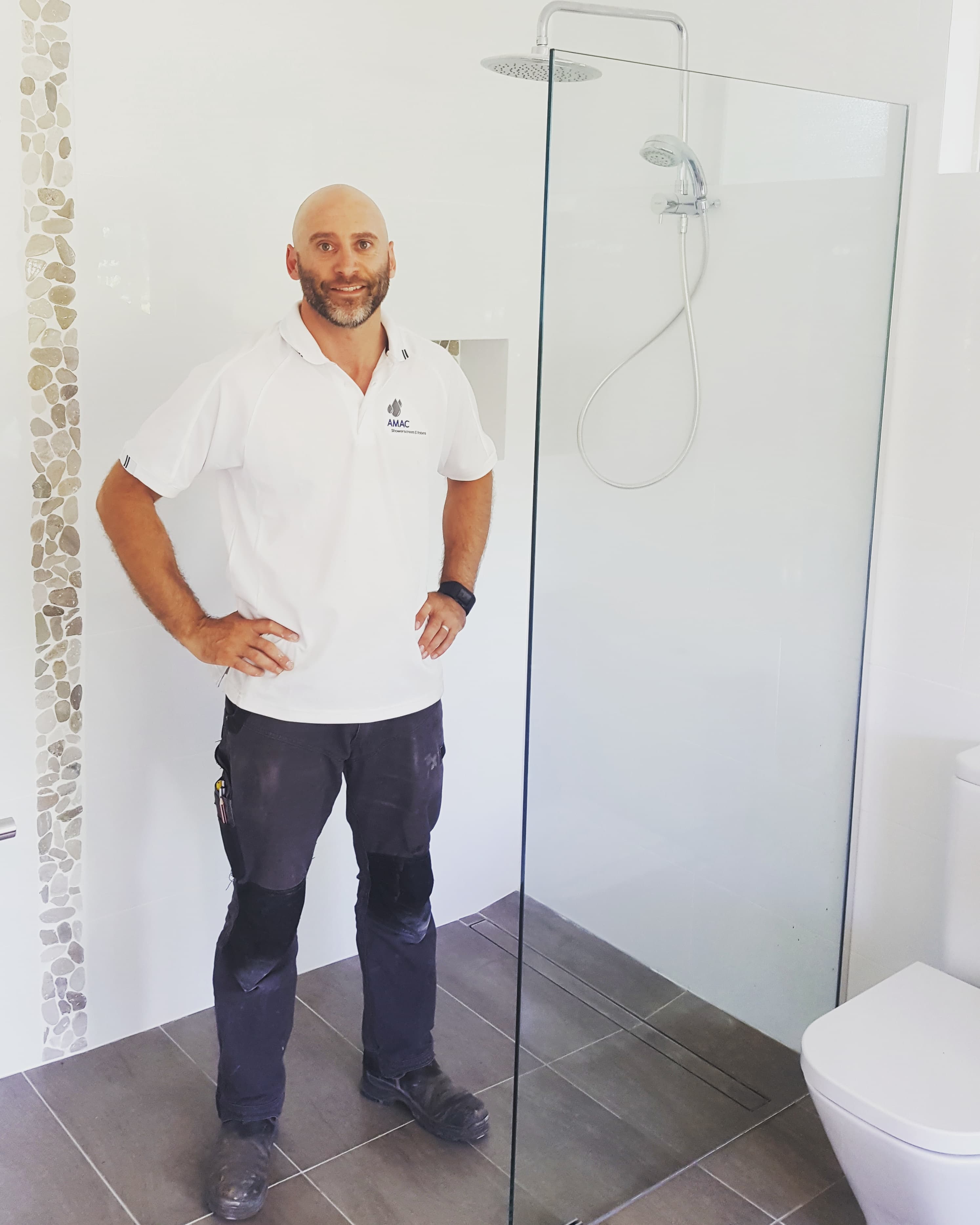 Smiling tradesman in white polo shirt standing beside a modern frameless glass shower screen.