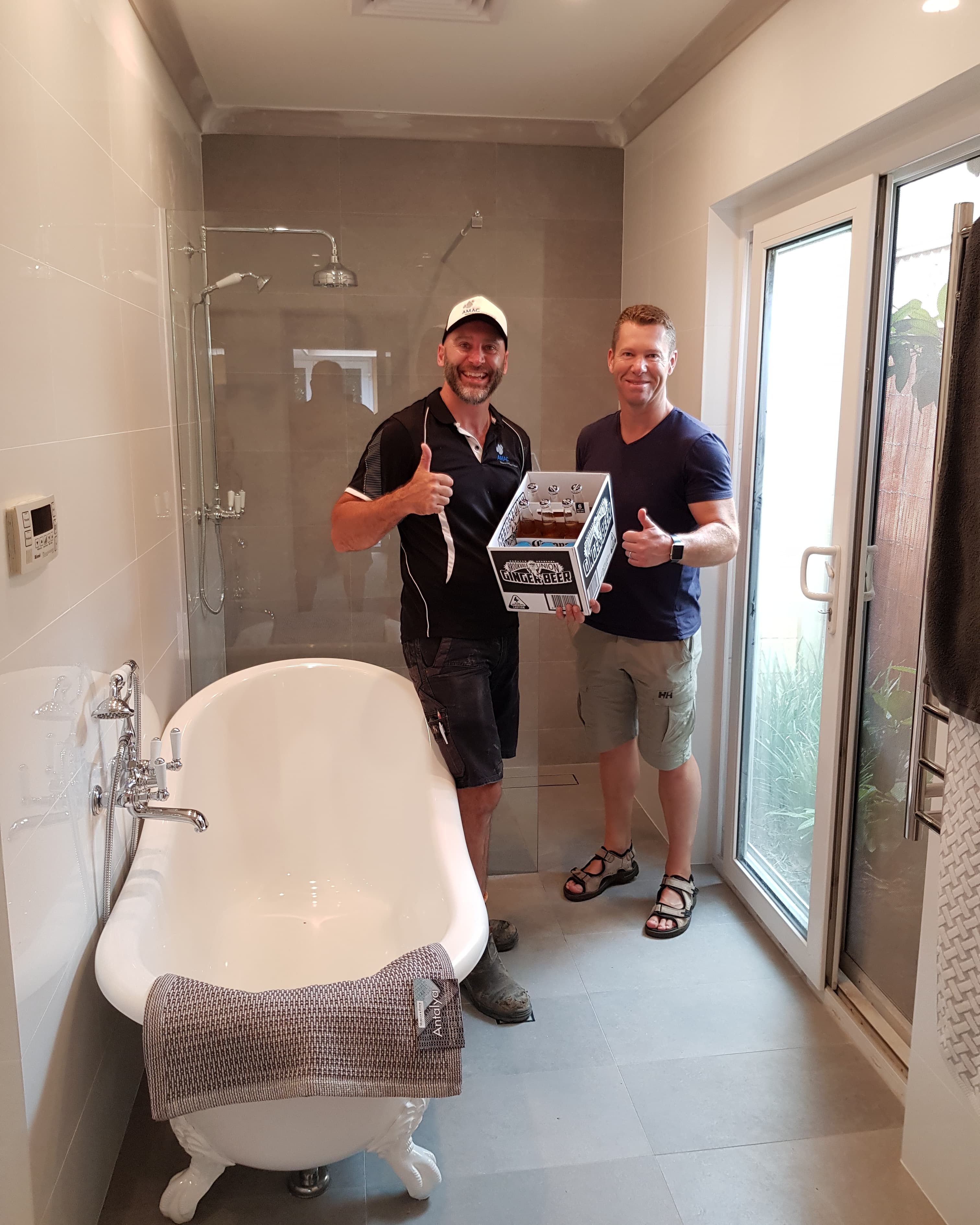 Two men giving thumbs up while holding ginger beer in a modern, renovated bathroom.