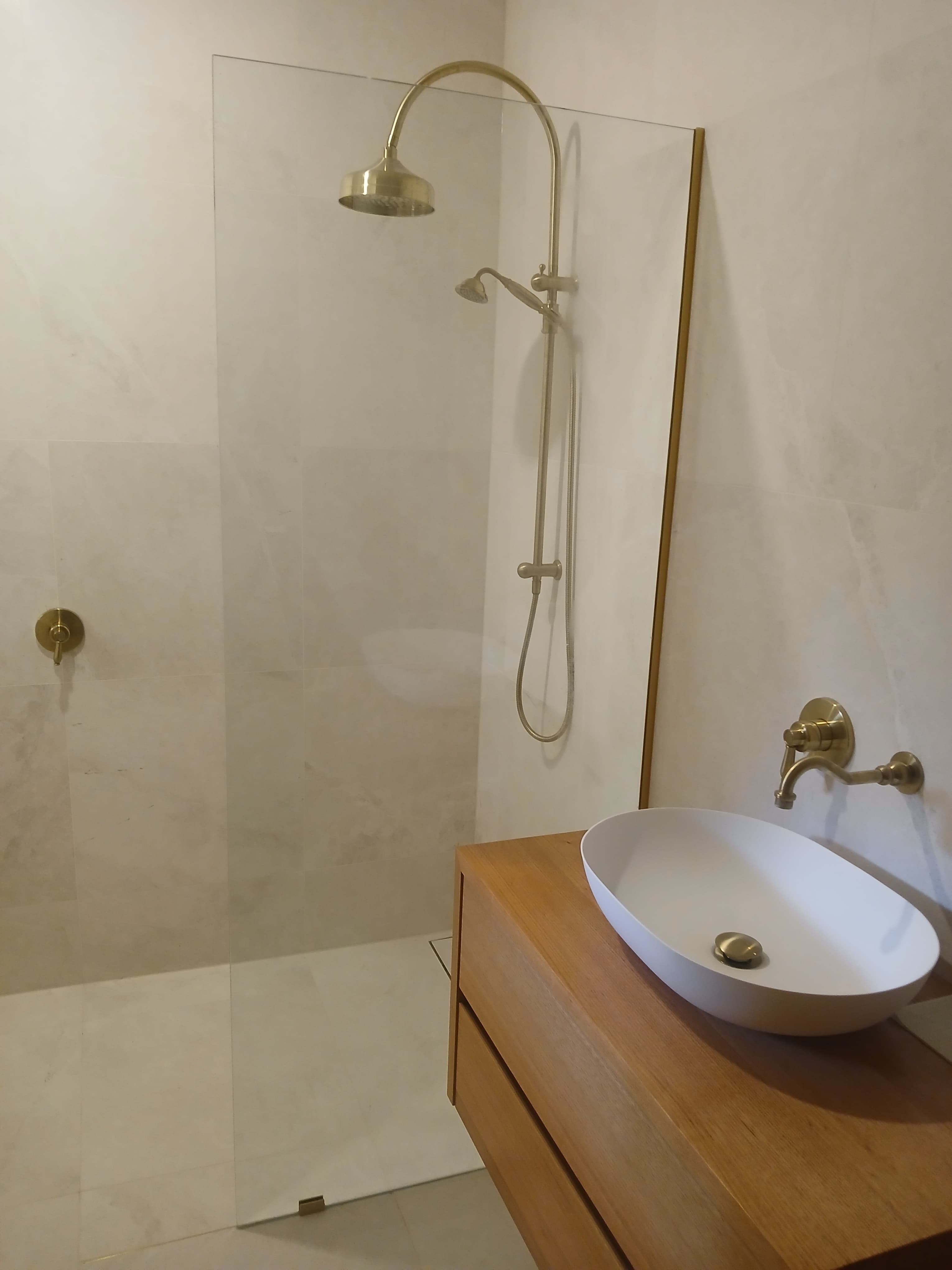 Modern bathroom with gold fixtures, glass shower partition, and wooden vanity with white vessel sink.