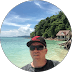 Man in sunglasses and cap at a tropical beach with turquoise water and lush hills.
